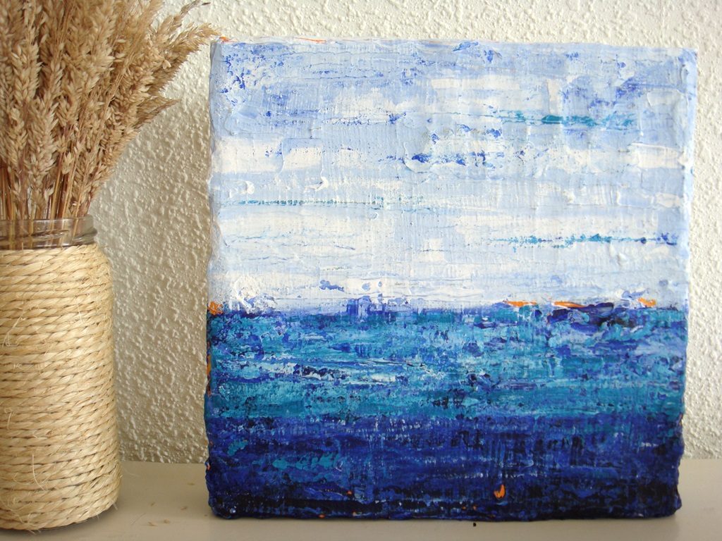 Schilderij lichtblauw - Kus van de zee II is een abstract klein schilderijtje met reliëf van 20 x 20 cm in (ultramarijn)blauw, turquoise, wit en oranje met stukjes juten, katoenen stof en zand erop.